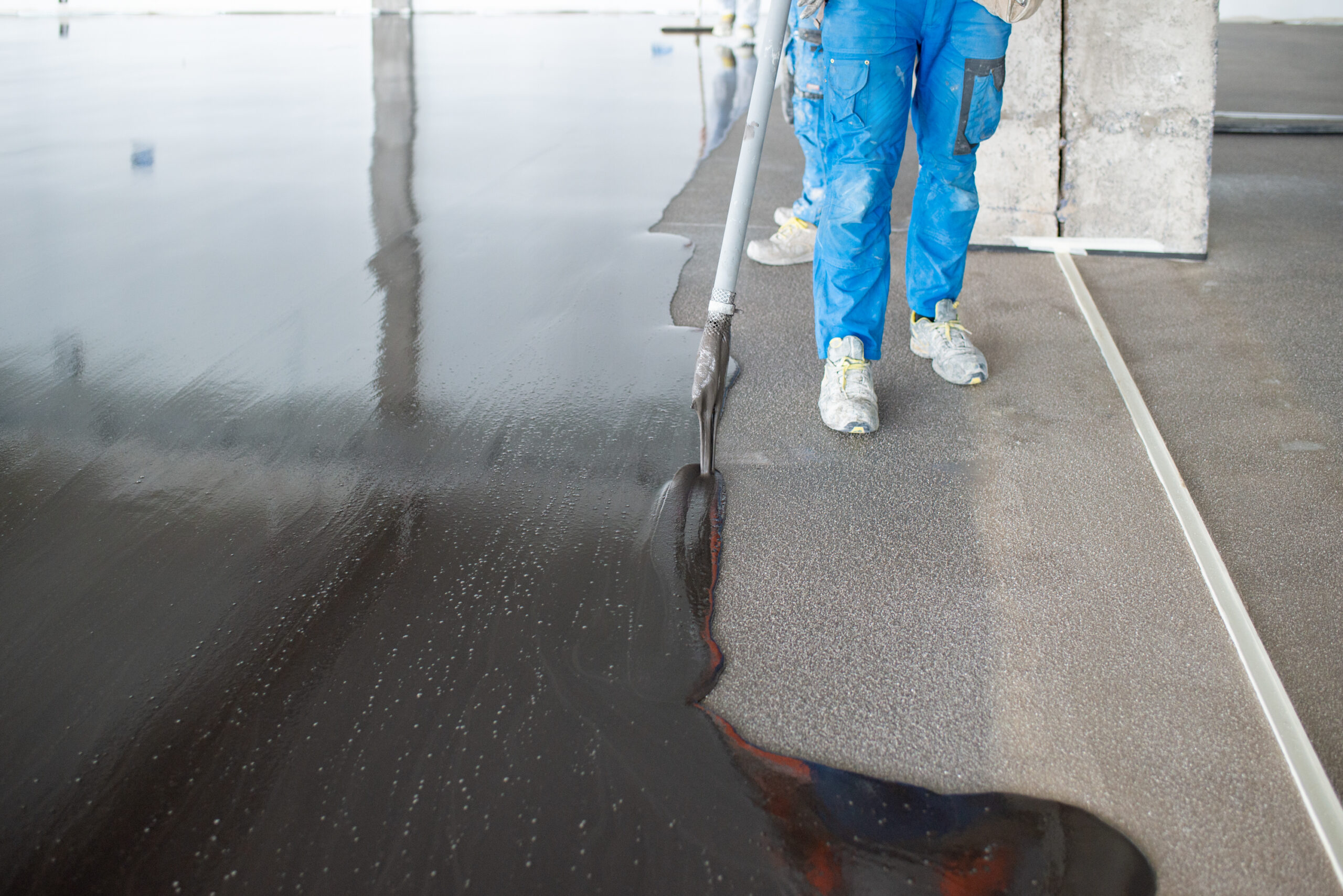 High angle of crop unrecognizable male workers in uniform and sneakers pouring and leveling liquid concrete on floor during renovation works at construction site